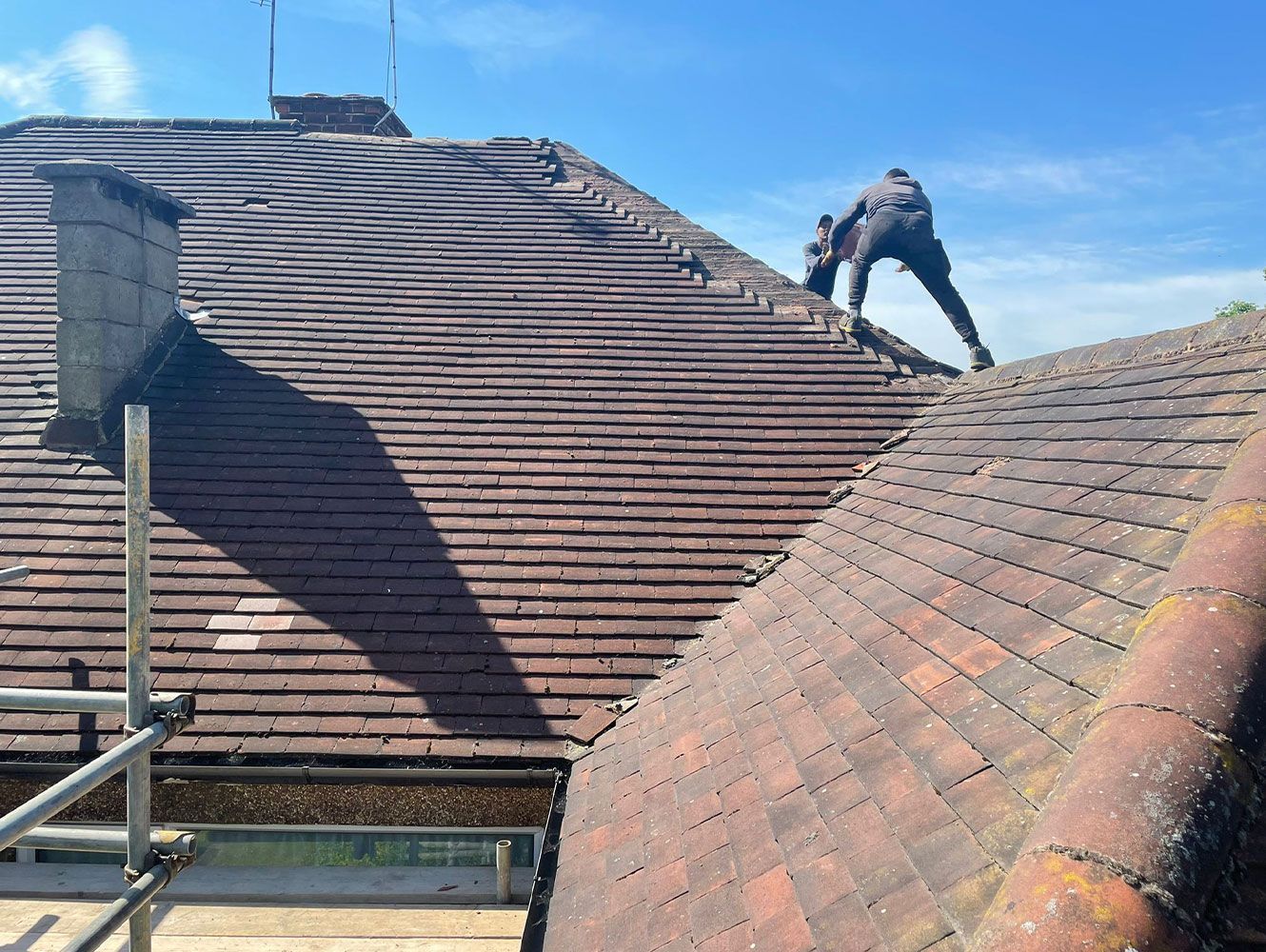 Roofer working on a weathered brown tile roof on a sunny day. Scaffolding visible.