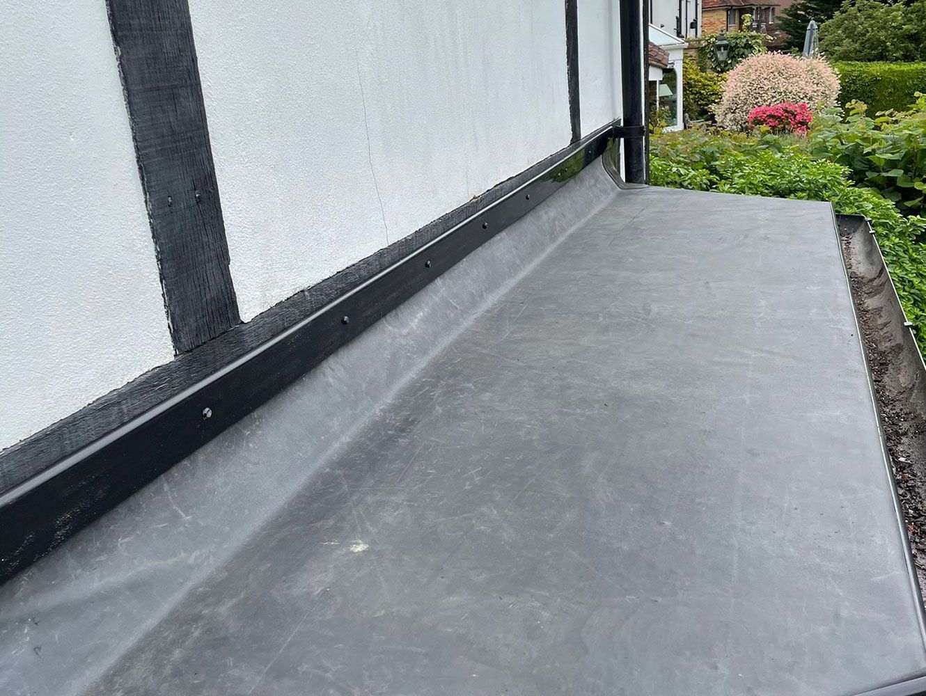 Black roofing material along a white wall with dark trim, with green foliage in the background.
