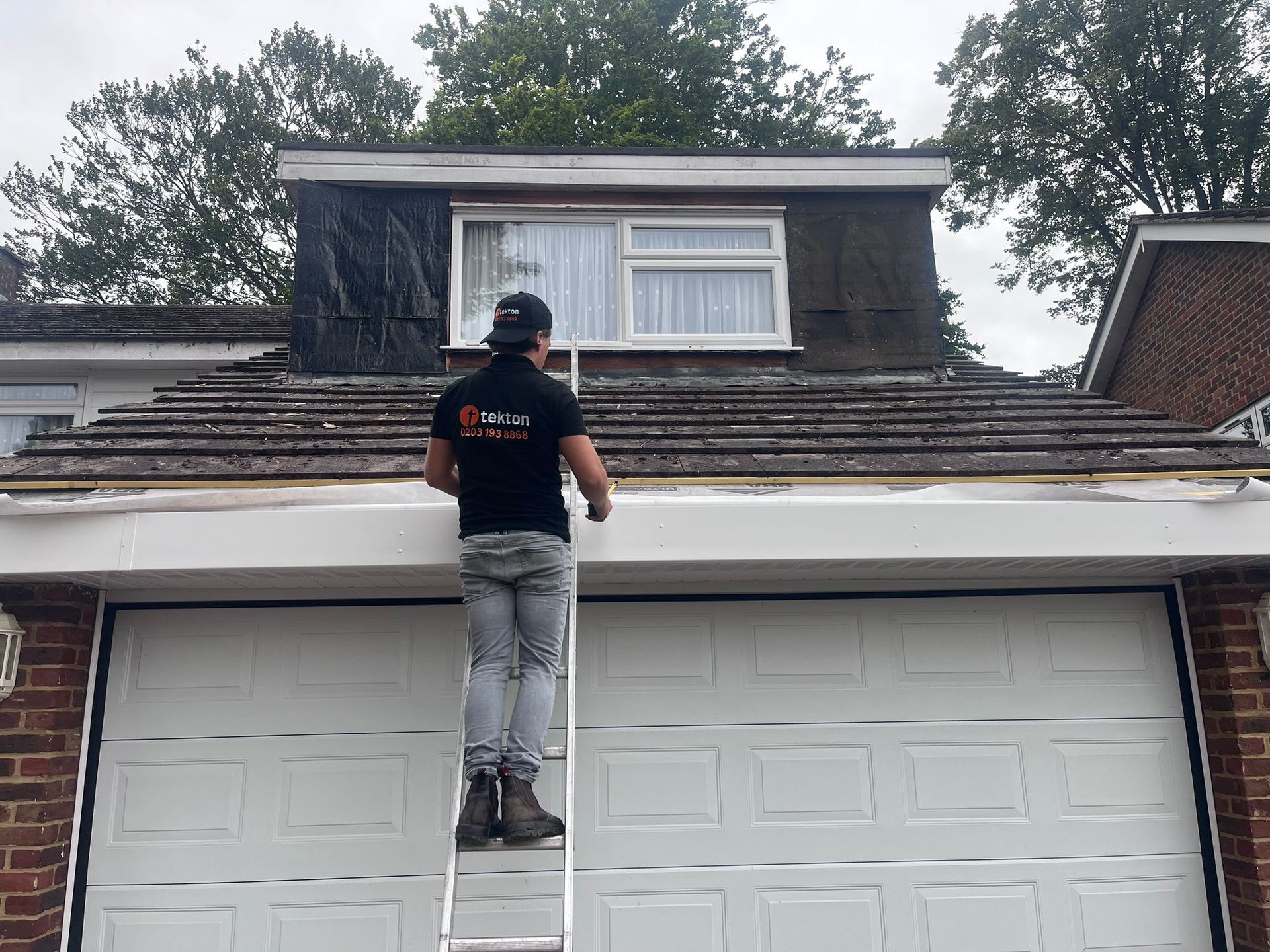 Person on ladder working on roof above a garage, overcast day.