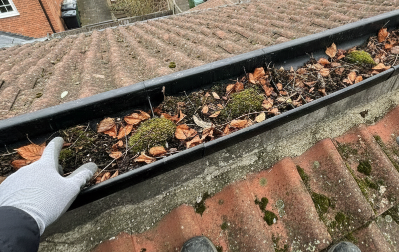 Gloved hand points to moss and leaves in a dirty rain gutter on a tiled roof.