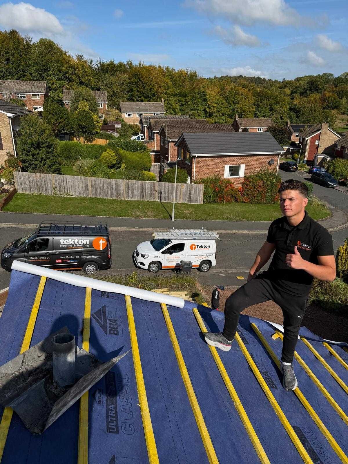 Man on rooftop giving thumbs up, two vans in background. Blue roof, sunny day, residential setting.
