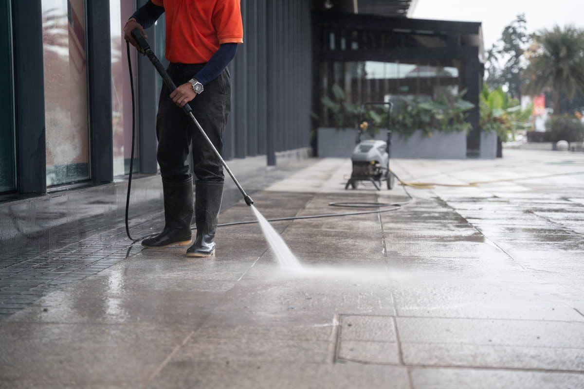 Person power washing a tiled sidewalk outside a building, using a pressure washer.