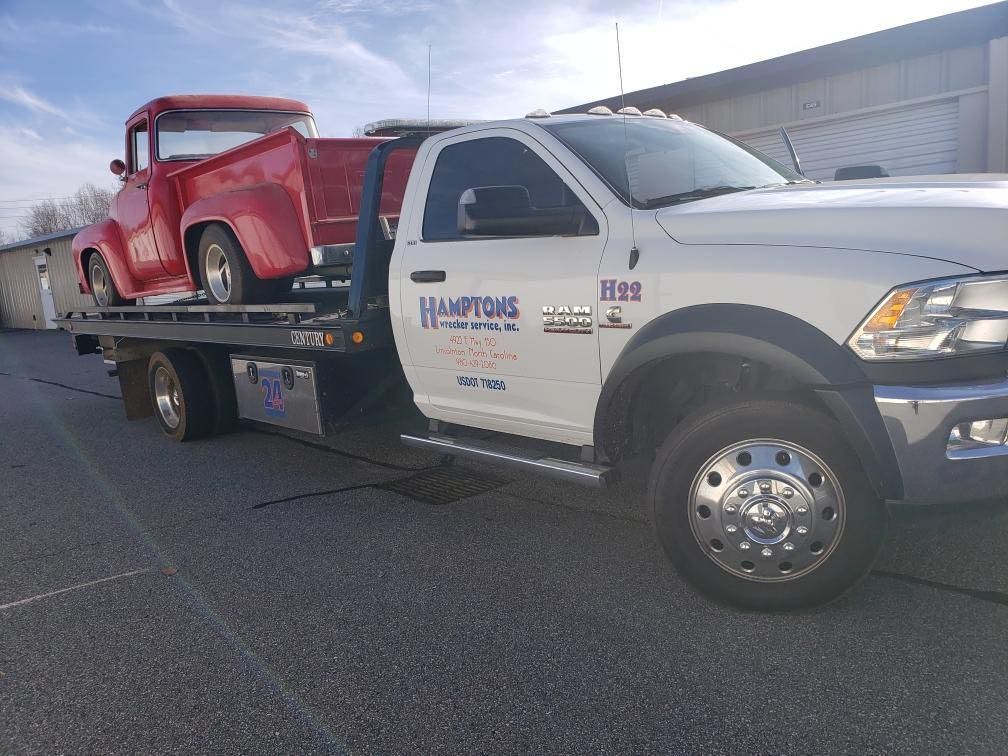 A red vintage pickup truck is being transported on a white Hampton's Towing flatbed truck in an outdoor setting.