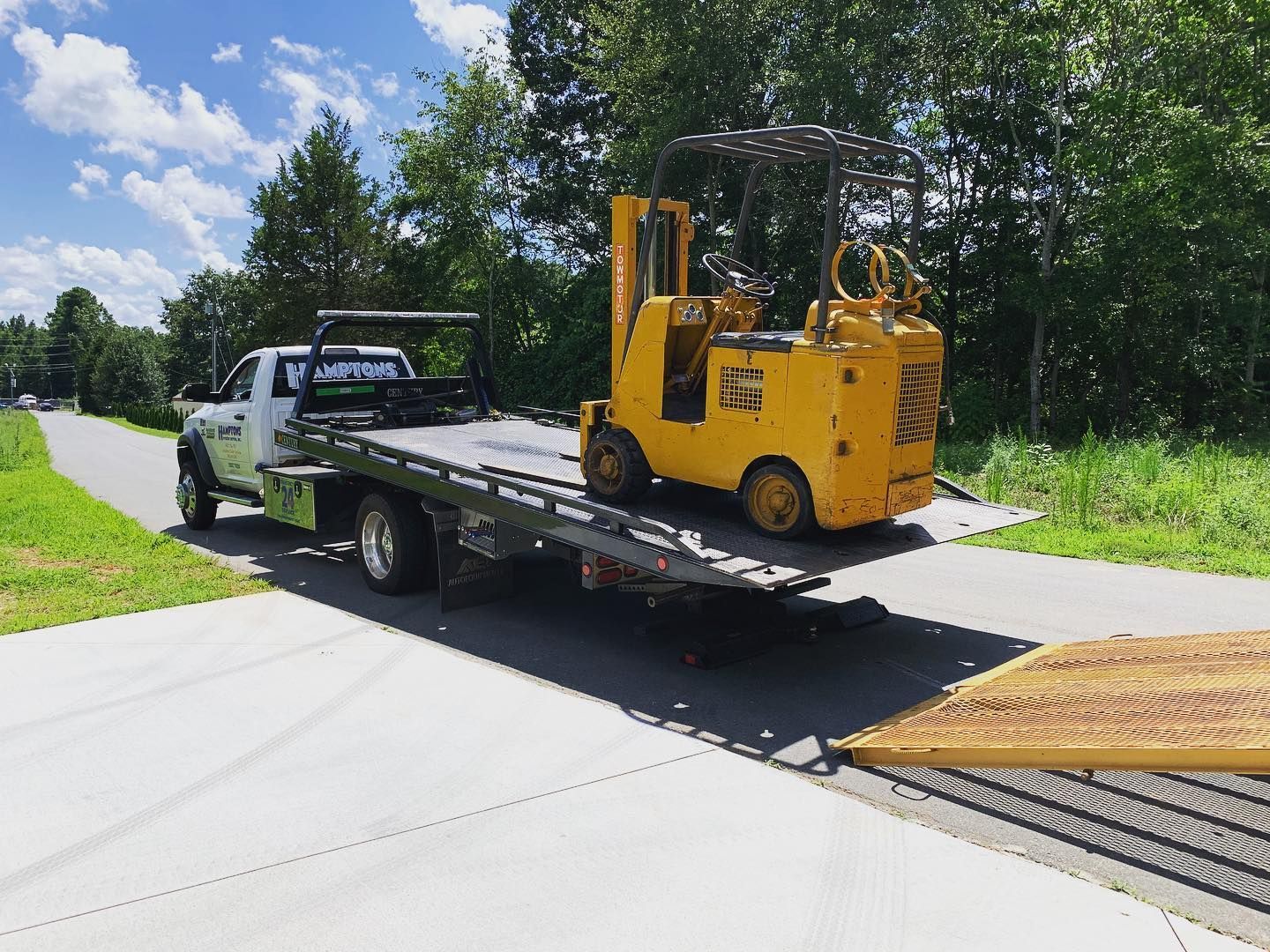 A yellow forklift is being towed on a flatbed tow truck on a paved road with trees in the background.