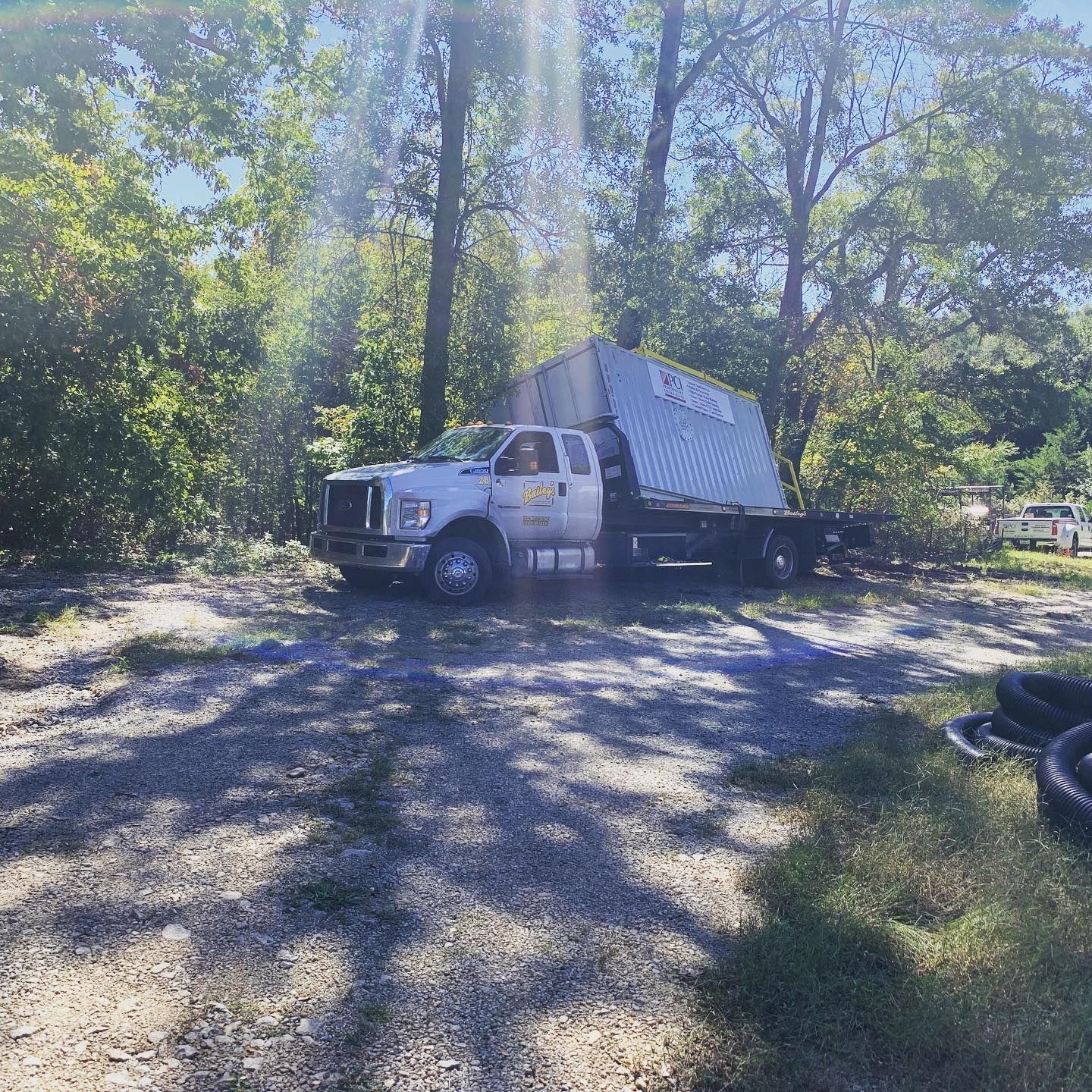 A white flatbed truck carrying a large, rectangular structure in a wooded area on a sunny day.