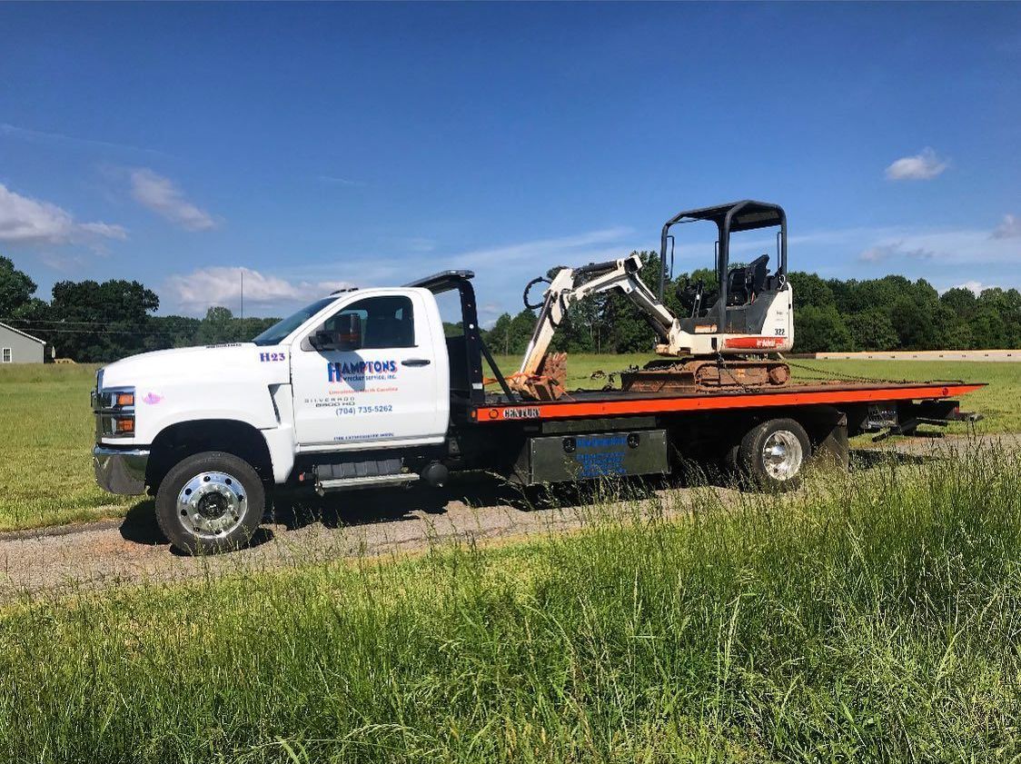 White flatbed tow truck with a Bobcat excavator on the back, parked on a grassy field under a blue sky.