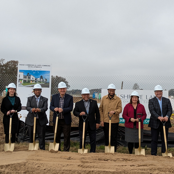Officials in hard hats hold shovels at a groundbreaking ceremony outdoors.