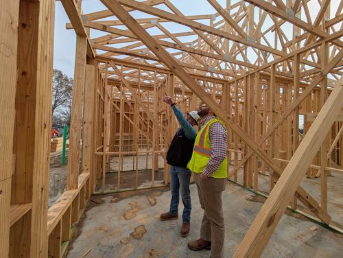 Two workers in safety vests inspect wooden house framing at a construction site.