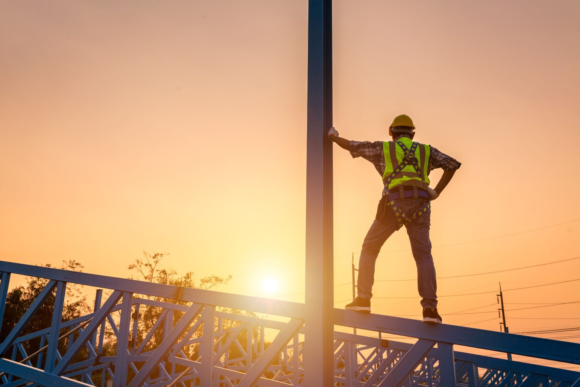 Construction worker standing on a steel beam at sunset, wearing a safety vest and helmet.