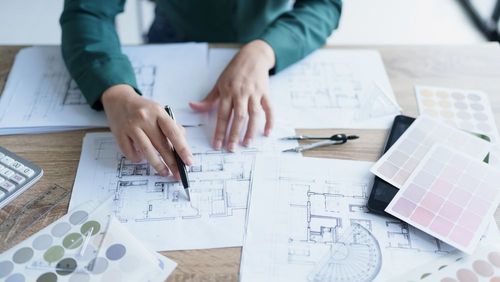 Hands sketching floor plans on architectural blueprints with pencils, ruler, and color swatches on a desk