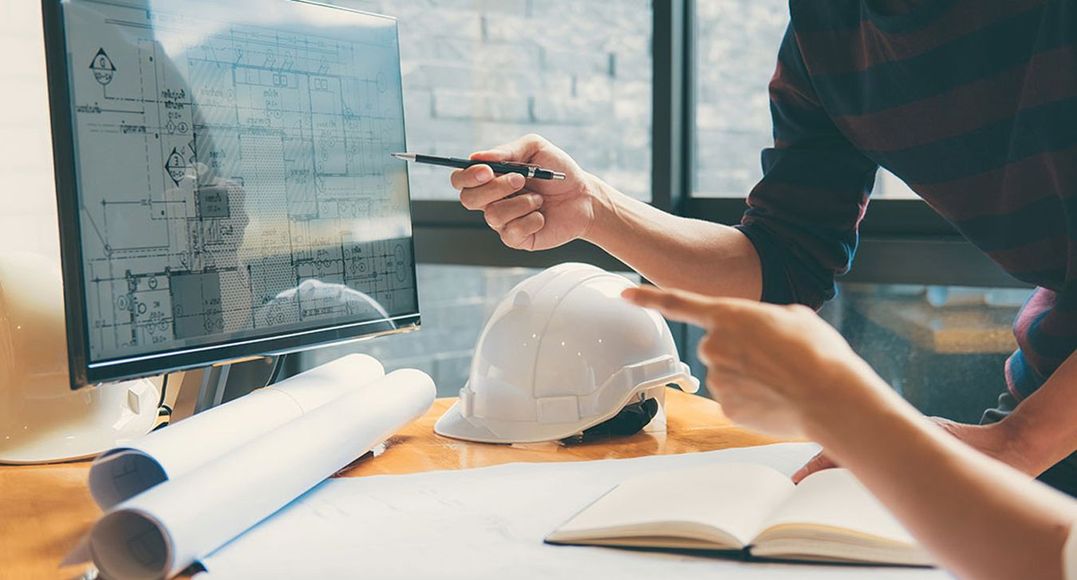 Engineer pointing at a computer monitor with blueprints beside a hard hat and open notebook on a desk