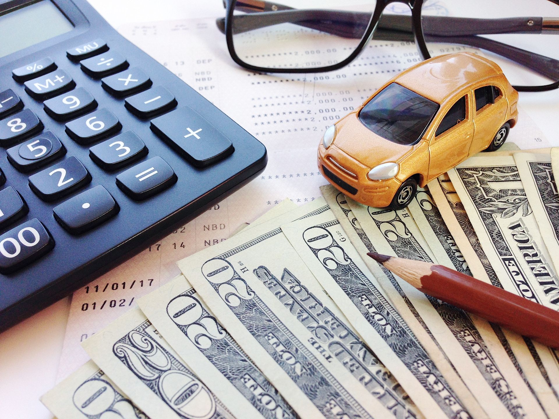 Calculator, dollar bills, toy car, glasses, and pencil on a desk.