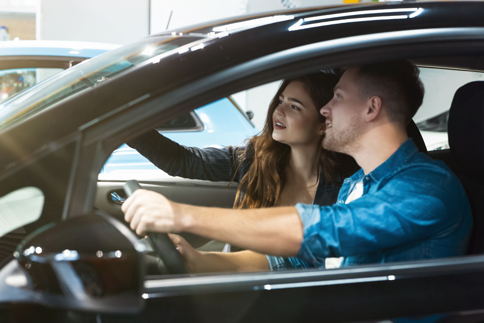 Two people in a car, one driving while the other points ahead and talks, inside a showroom-like setting