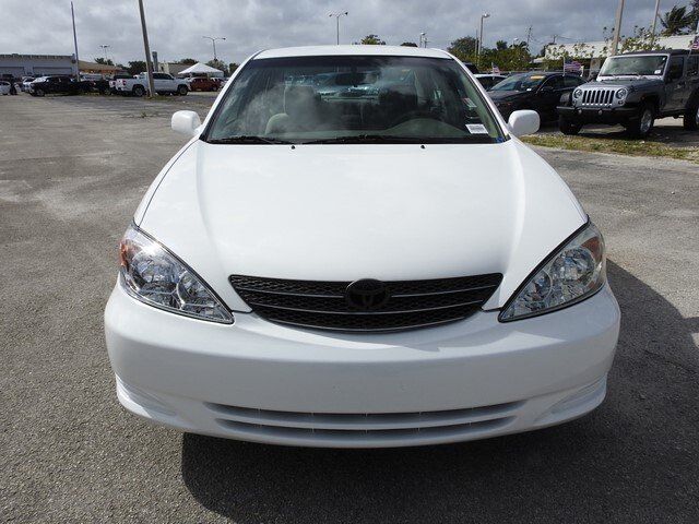A white Toyota Camry parked in an outdoor car lot under a partly cloudy sky.