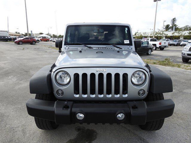 A silver Jeep Wrangler parked on a gravel lot under a bright, overcast sky.