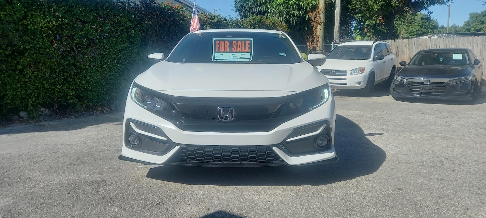 A white Honda Civic parked on a gravel lot with a "For Sale" sign in the windshield.