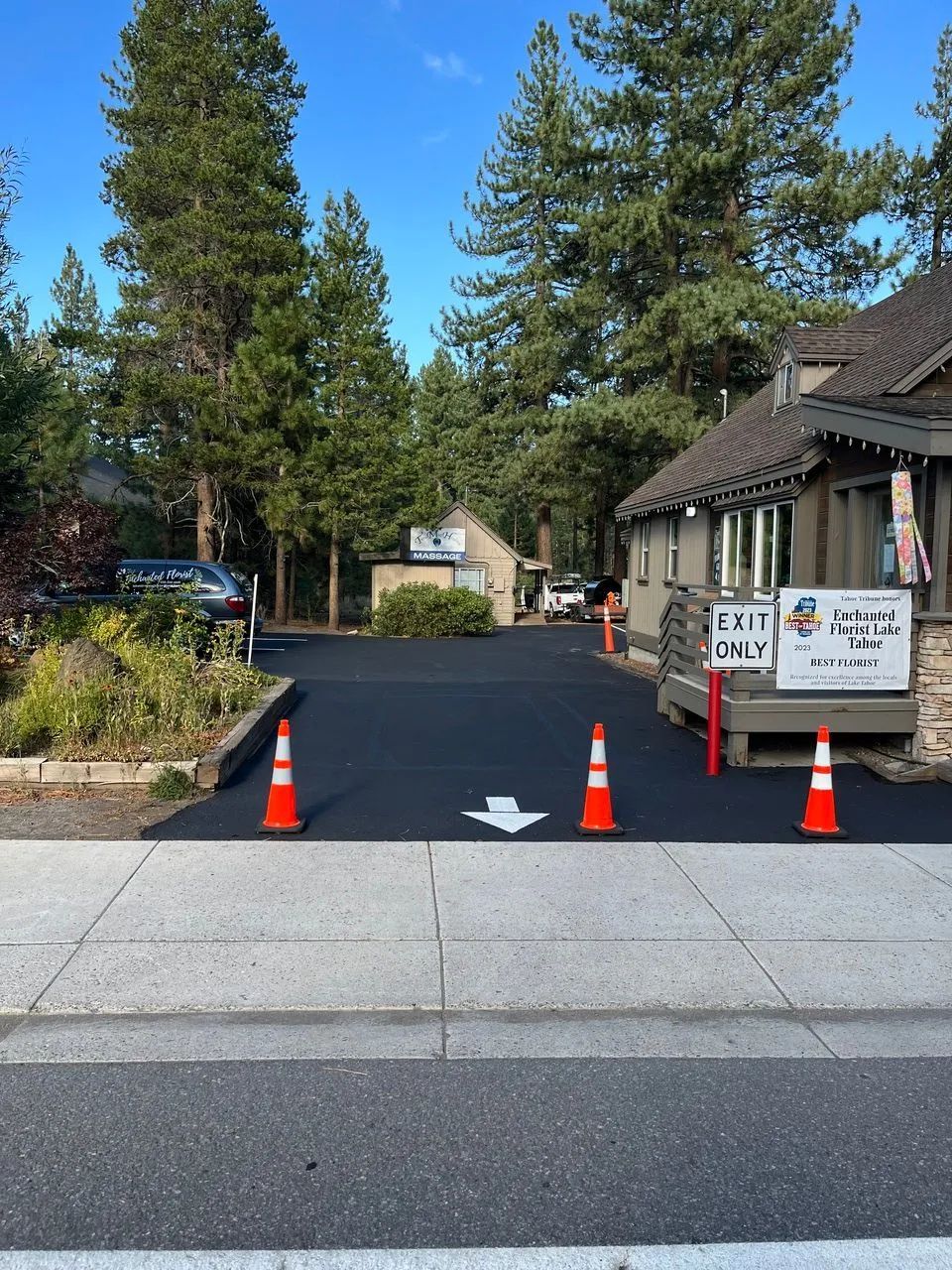 Driveway entrance blocked by orange cones and a blacktop barrier next to a building and sign.