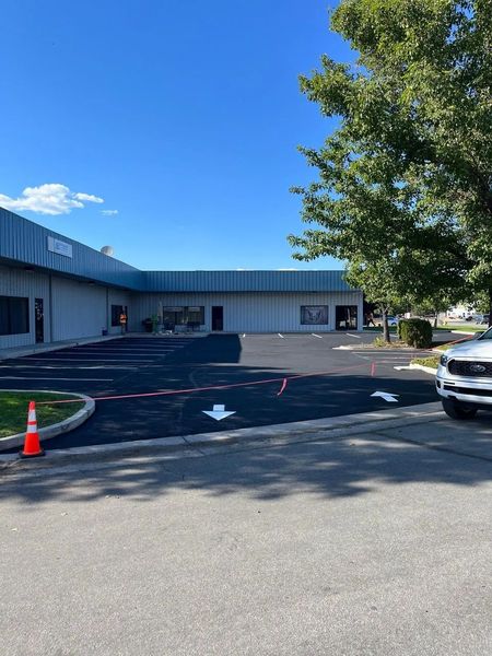 Empty parking lot beside a low building under a bright blue sky, with trees and a parked car.