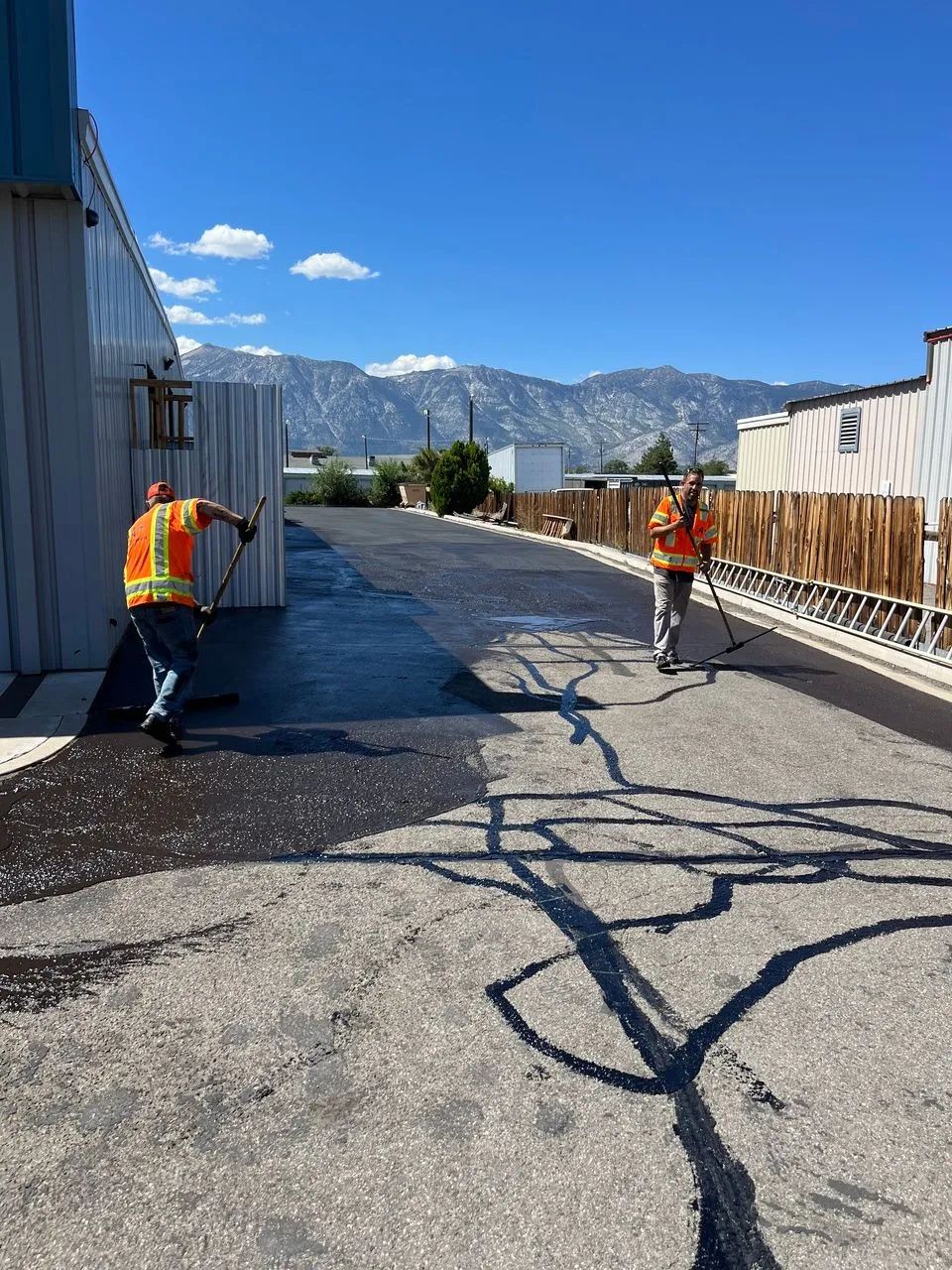 Two road workers in orange vests repaving a cracked street with mountains in the background