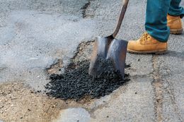 Worker shoveling black asphalt mix into a pothole on a cracked road surface