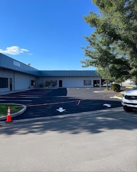 Small parking lot beside a blue-gray building, with orange cones, road arrows, and a tree by a parked car.