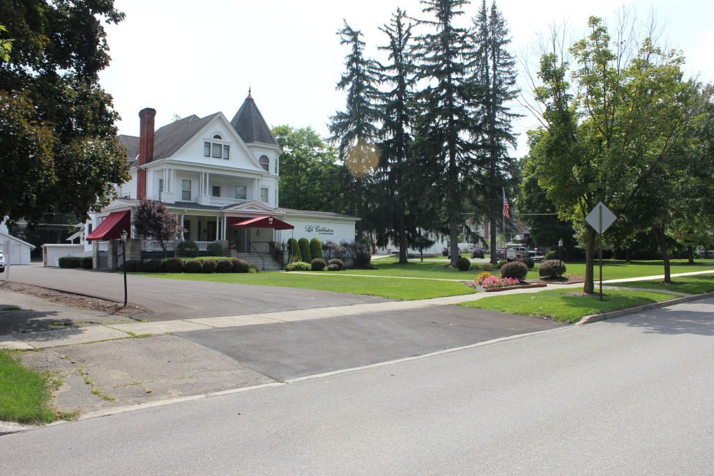 Large white Victorian house with a red awning, set back from a street with green lawns and trees.
