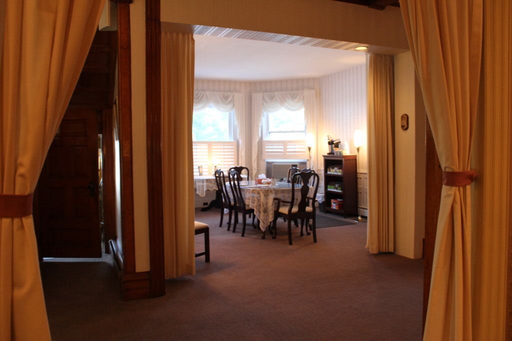 Interior view of a dining room, seen through an archway with tan curtains. A table is set with chairs, soft light, and wallpapered walls.