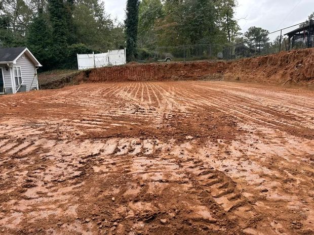 Cleared muddy construction lot with tire tracks, a small shed on the left, and trees in the background