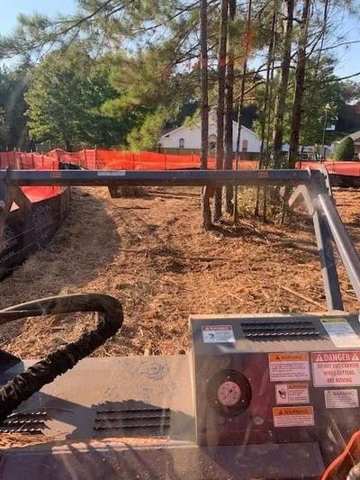 Construction machinery clearing dirt along an orange safety fence in a wooded area, with a white house in the background