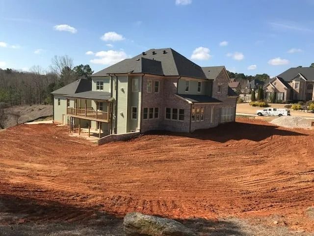 Large brick house under construction on a red dirt lot with a partially finished roof and clear blue sky.