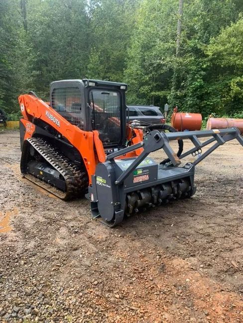 Orange and black skid steer with grapple attachment on muddy ground near a wooded area