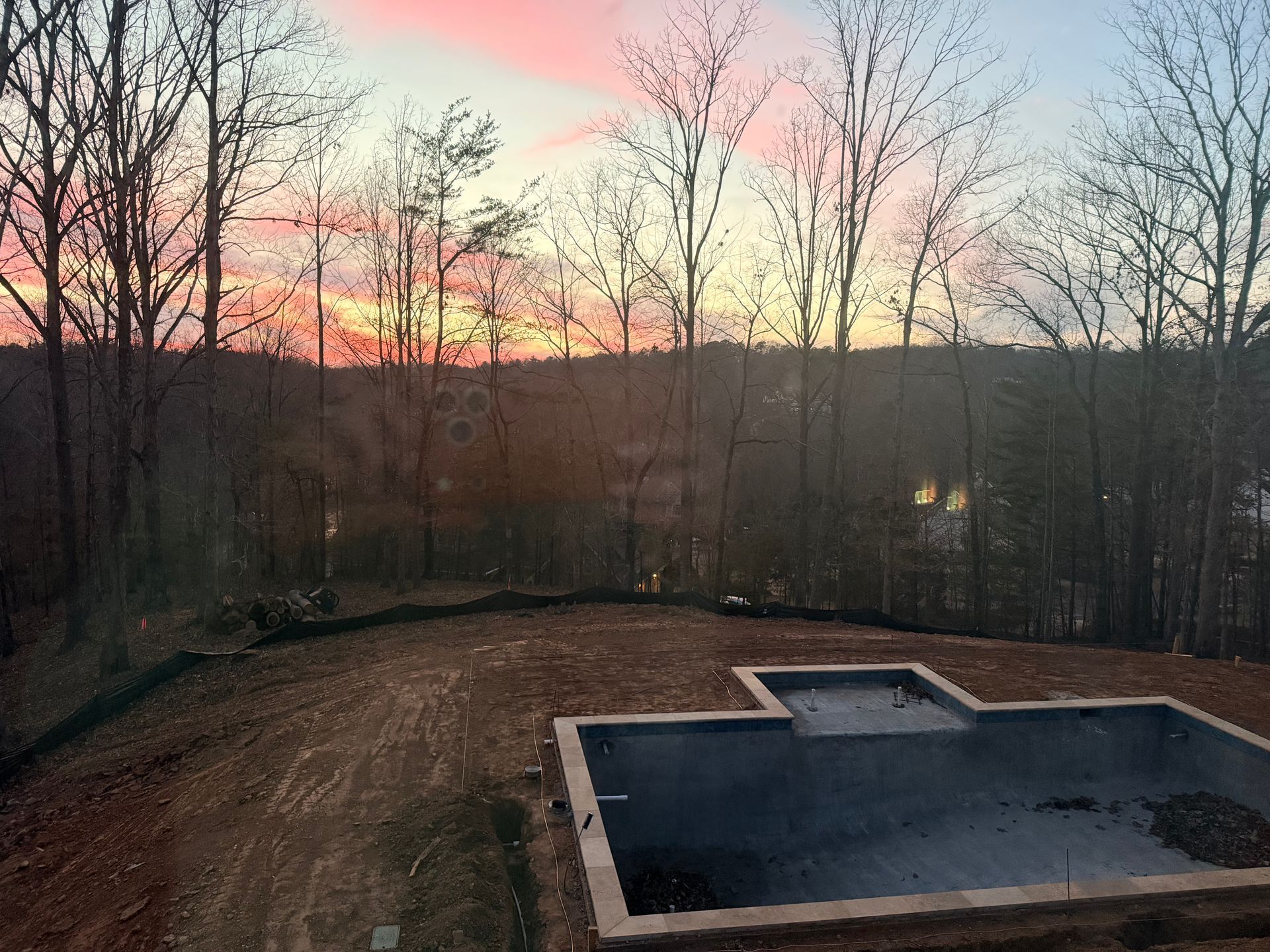Backyard pool at dusk with bare trees and a pink-orange sunset sky