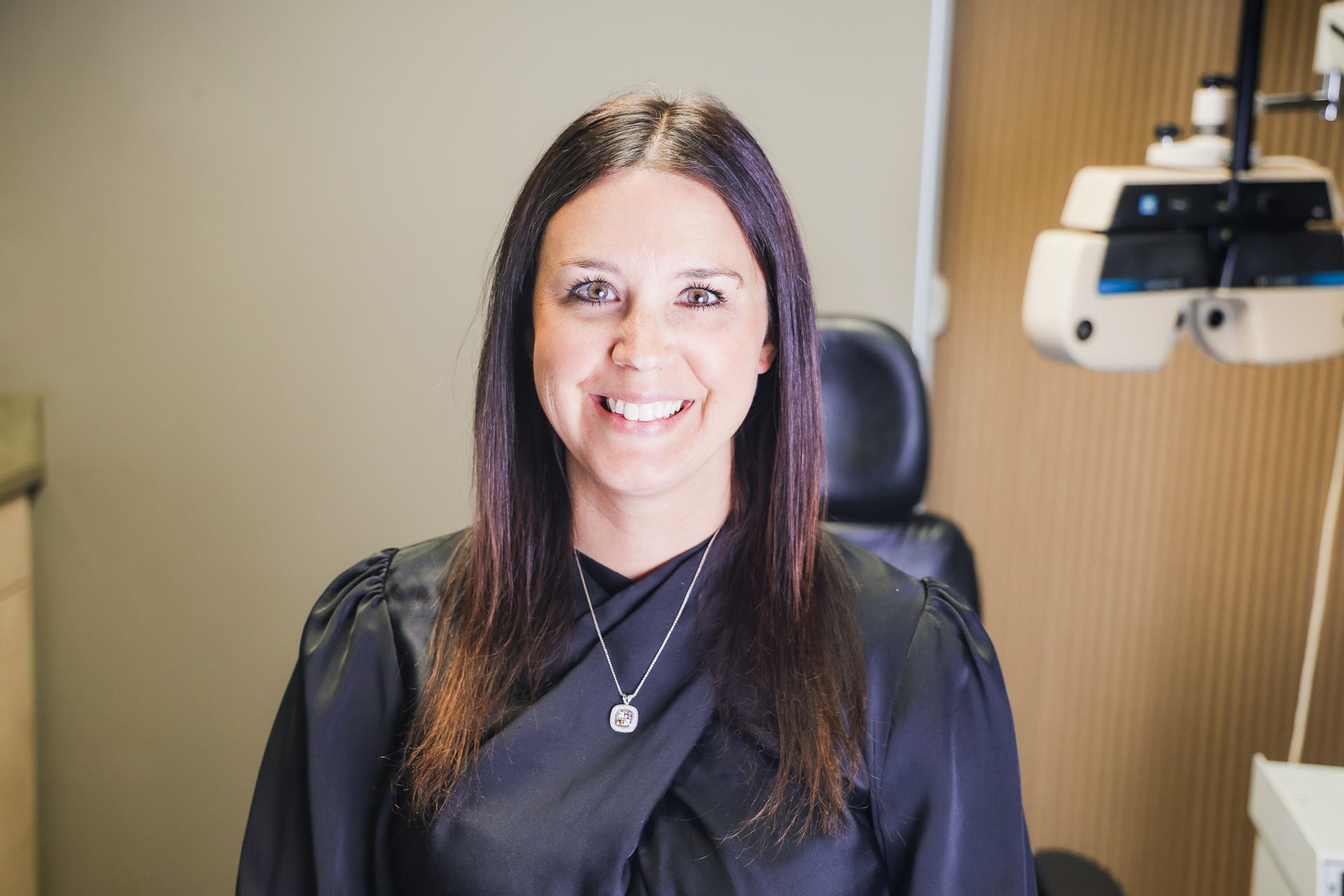 Smiling woman with long dark hair in a teal top, standing in a bright showroom with eyeglass frames