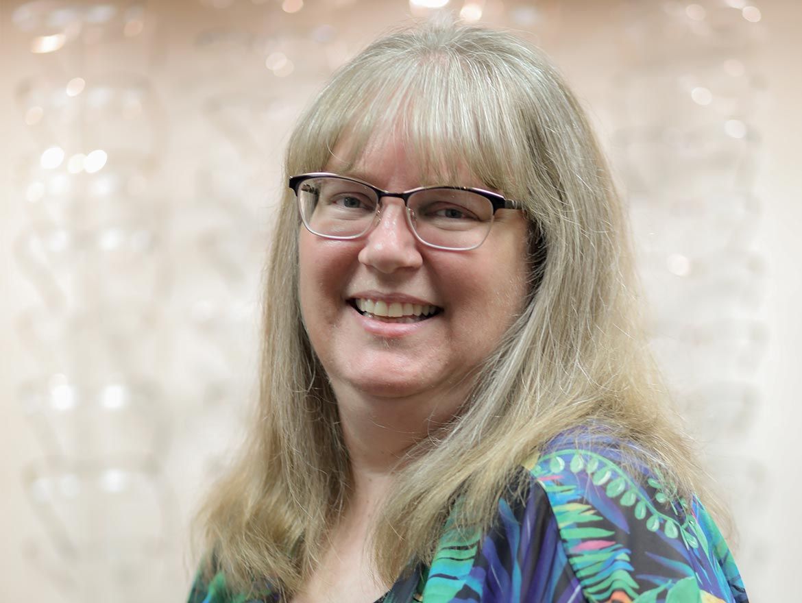 Smiling woman with long dark hair in a teal top, standing in a bright showroom with eyeglass frames