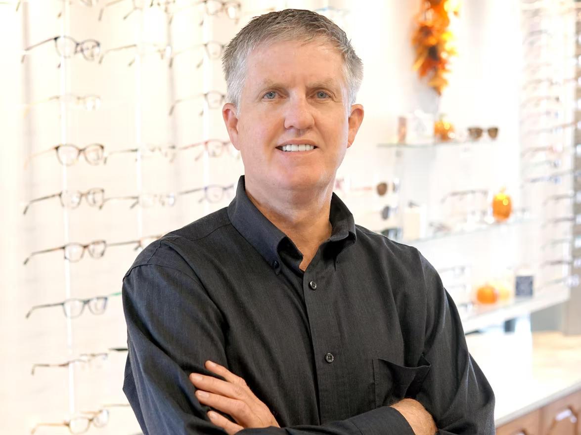 Smiling man in a dark shirt standing with arms crossed in an eyeglasses shop