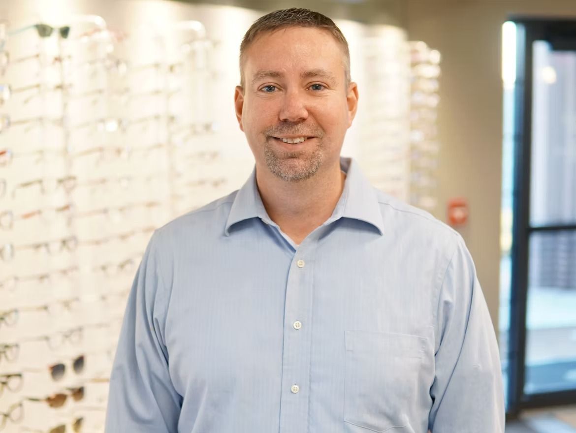 Smiling man in a dark shirt standing with arms crossed in an eyeglasses shop