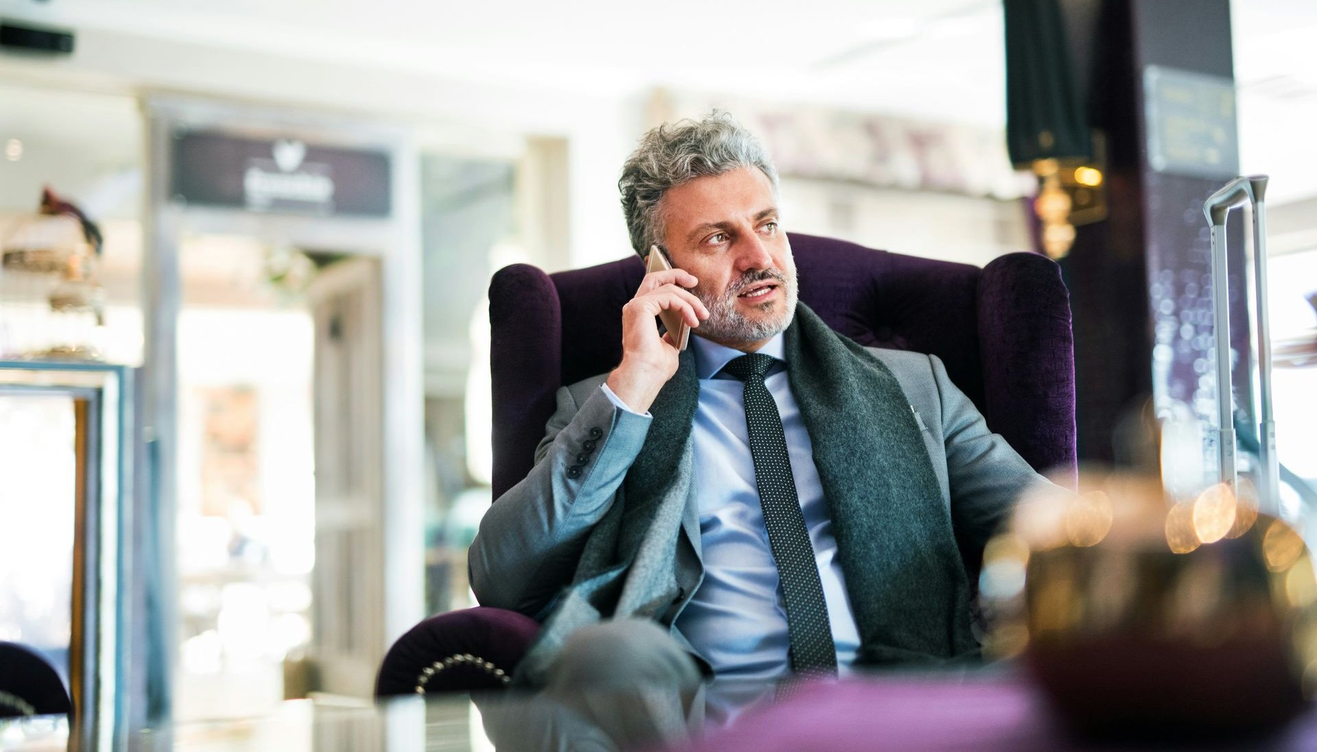 A person in a suit and scarf sits in a purple armchair while talking on a smartphone.
