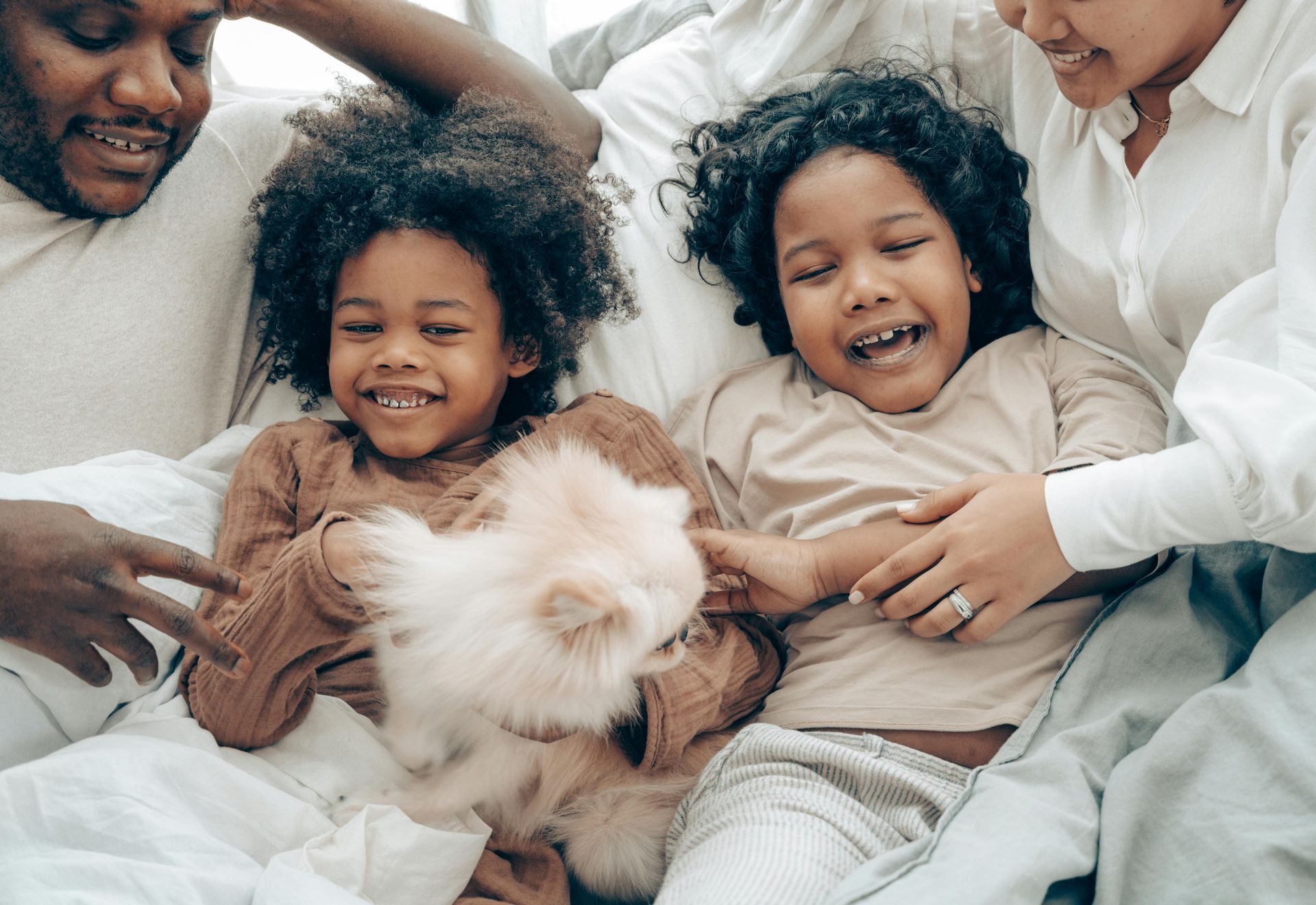 A family of four lying together on a bed, smiling while playing with a fluffy white pet.