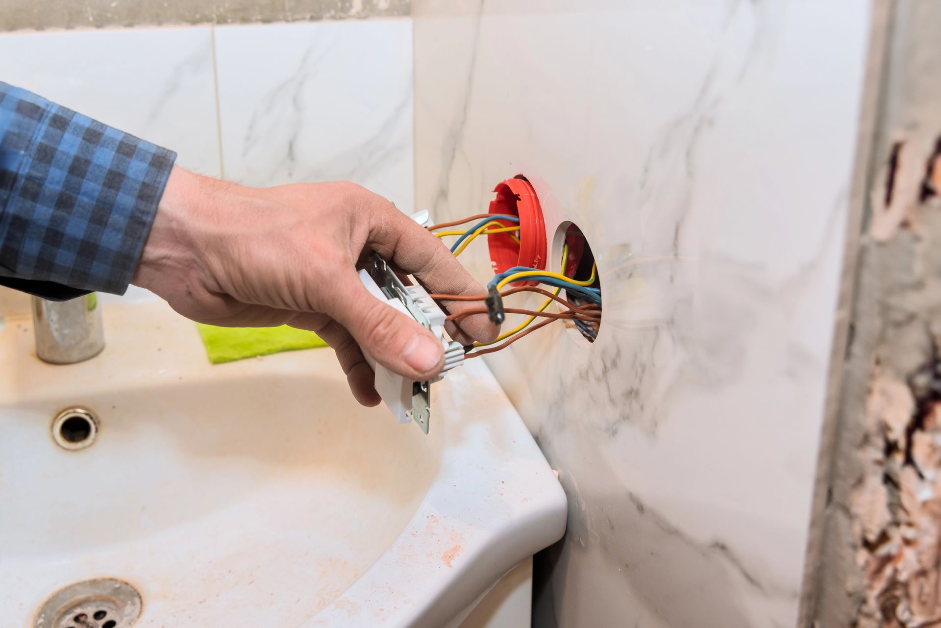 Mano che regge dei fili elettrici scoperti vicino a un lavandino in una parete piastrellata del bagno.