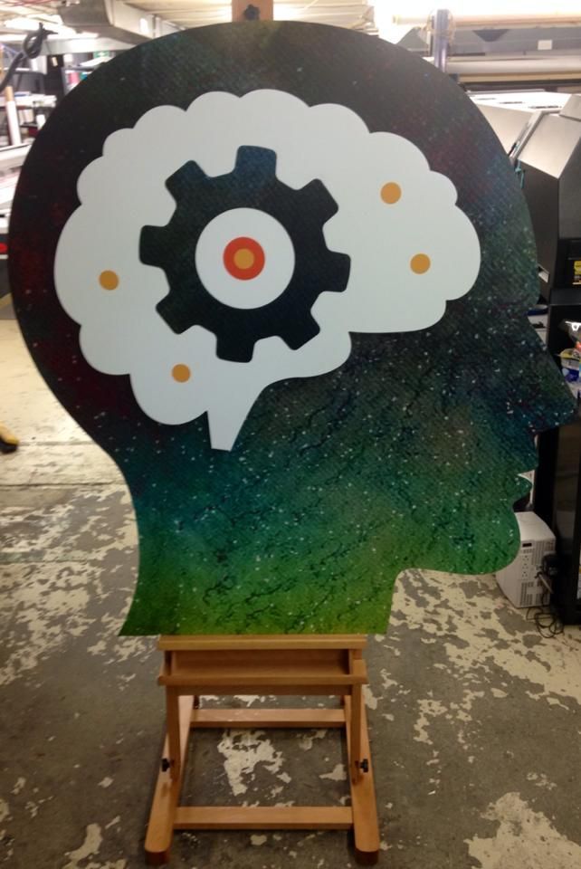 Silhouette head artwork with a white gear cloud inside, painted in black, blue, green, and orange on an easel.