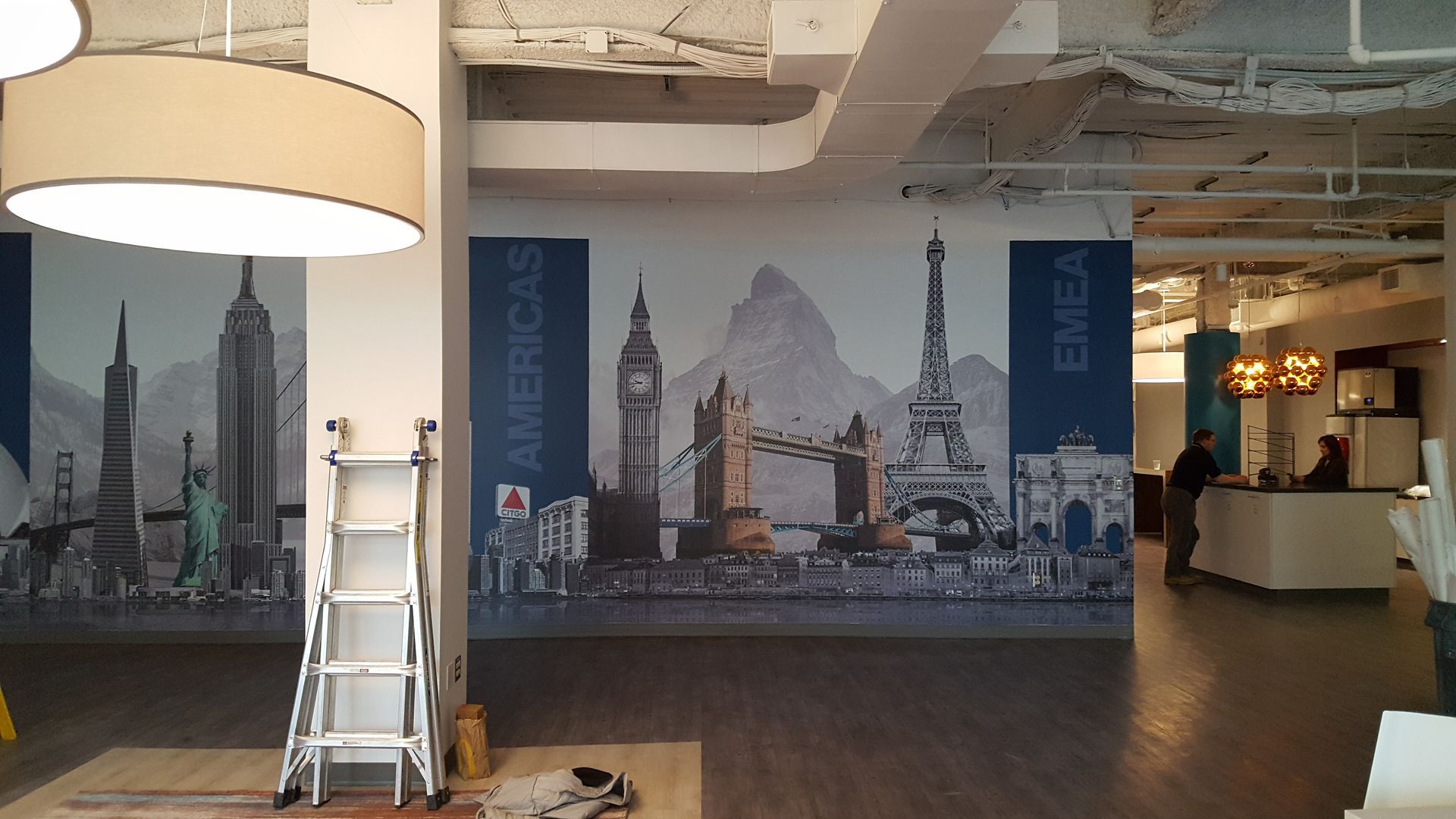 Modern office lobby with a large black-and-white cityscape mural, ladder, and hanging lamp.