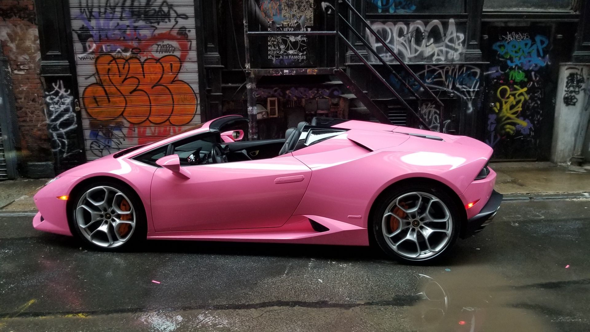 Pink Lamborghini convertible parked on a wet, graffiti-covered street.