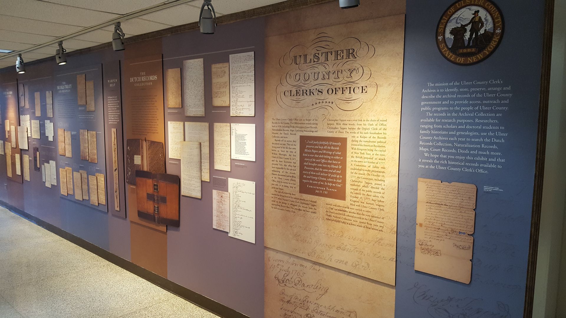 Museum hallway wall display of historical panels, documents, and plaques in blue and tan tones