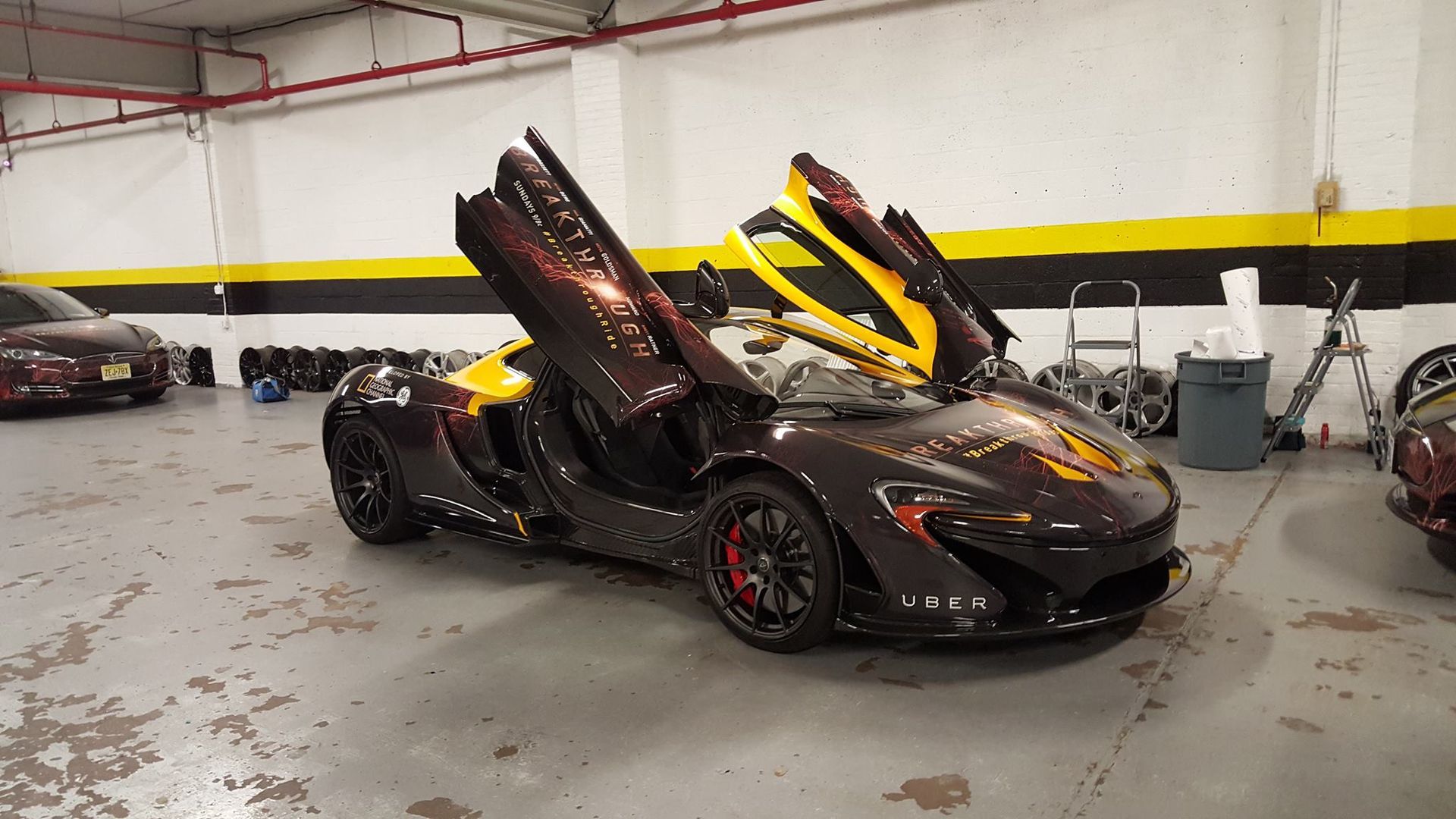 Black McLaren P1 sports car with butterfly doors open in a garage, yellow and red accents.
