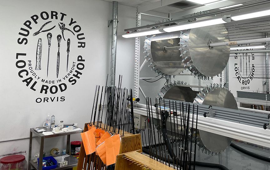 Orvis rod shop interior with tools, rods, and rotating drying wheels; "Support Your Local Rod Shop" text on the wall.