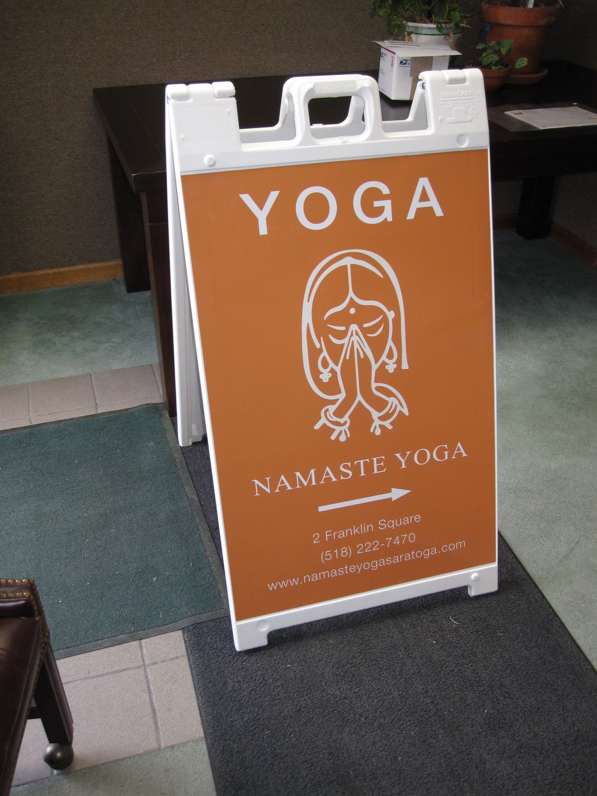 Orange sidewalk sign advertising yoga classes with “YOGA” and a seated figure icon.