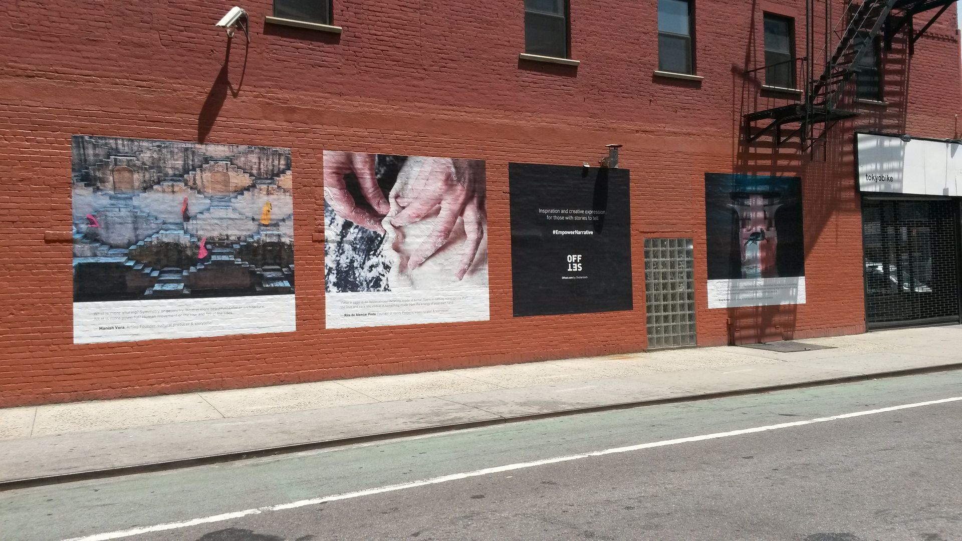 Brick wall with four large posters along a city sidewalk on a sunny street.
