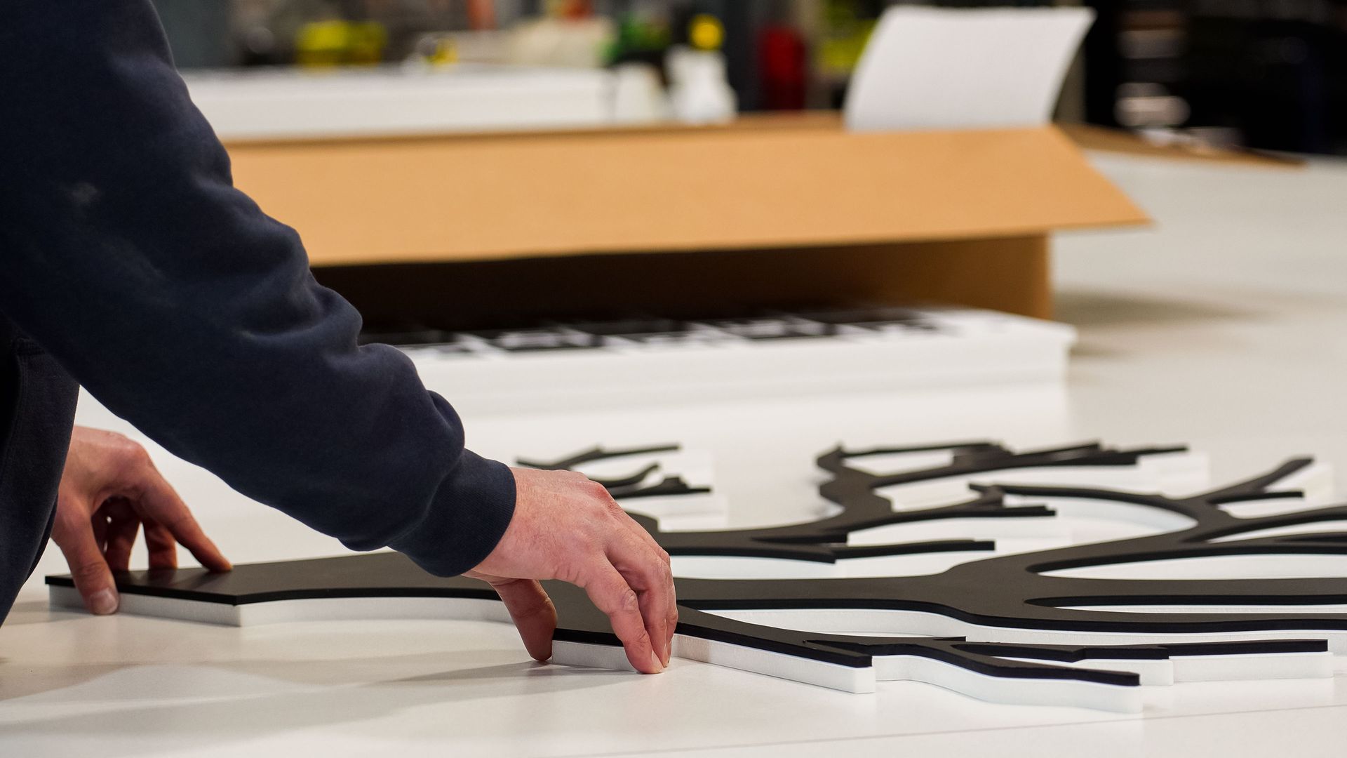 Person's hands assembling a black tree-shaped sign on a white surface, with a cardboard box in the background.