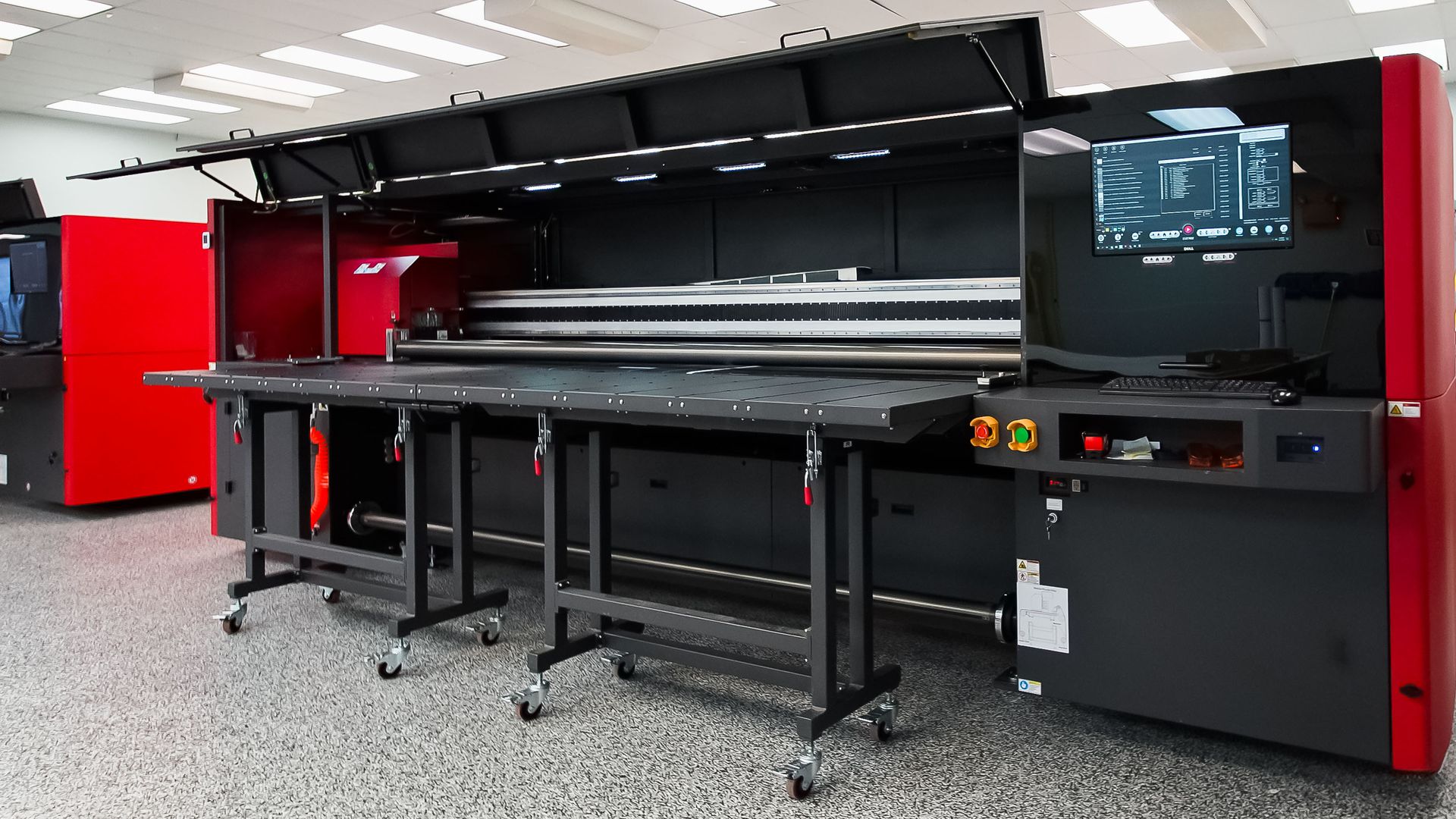 Large, industrial flatbed printer with black and red accents. Features a long, flat printing surface on a wheeled stand.