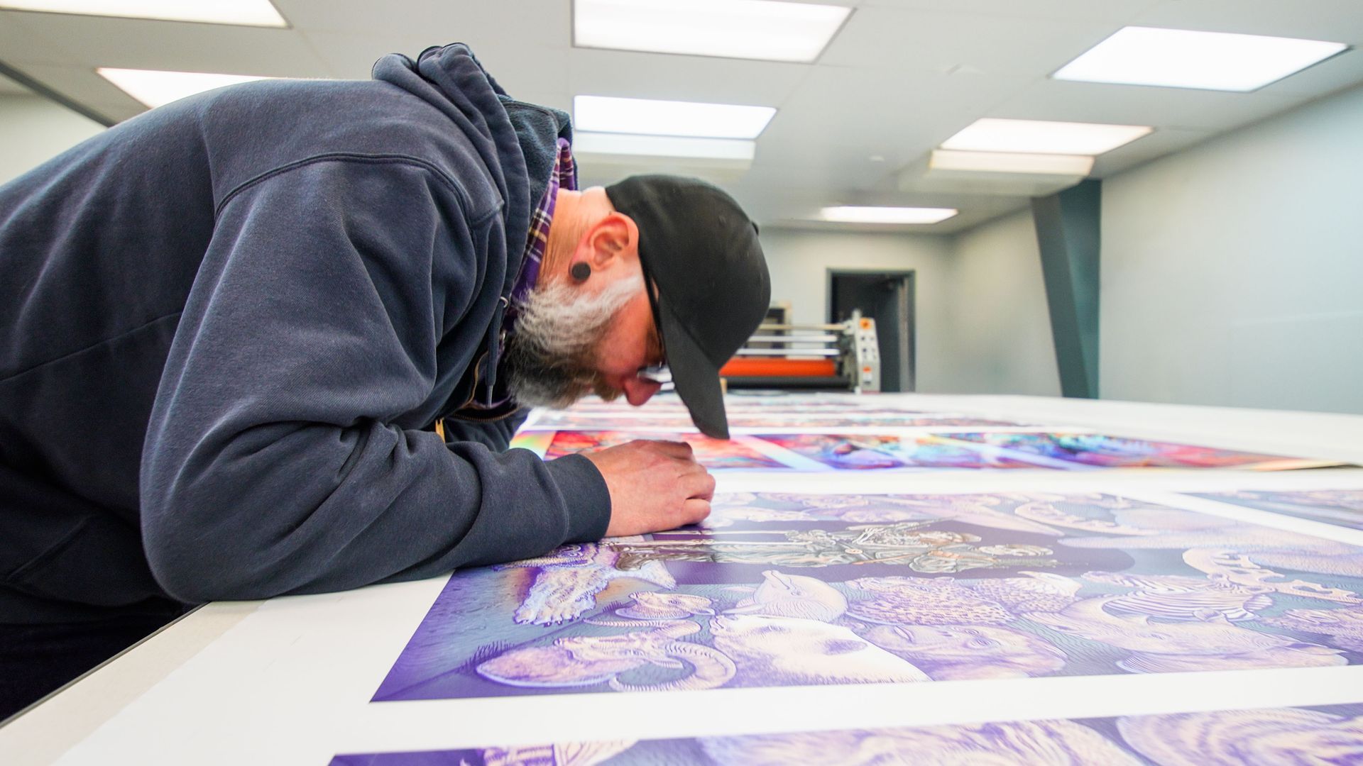 Person in a cap leans over a large print, carefully drawing in a bright studio.